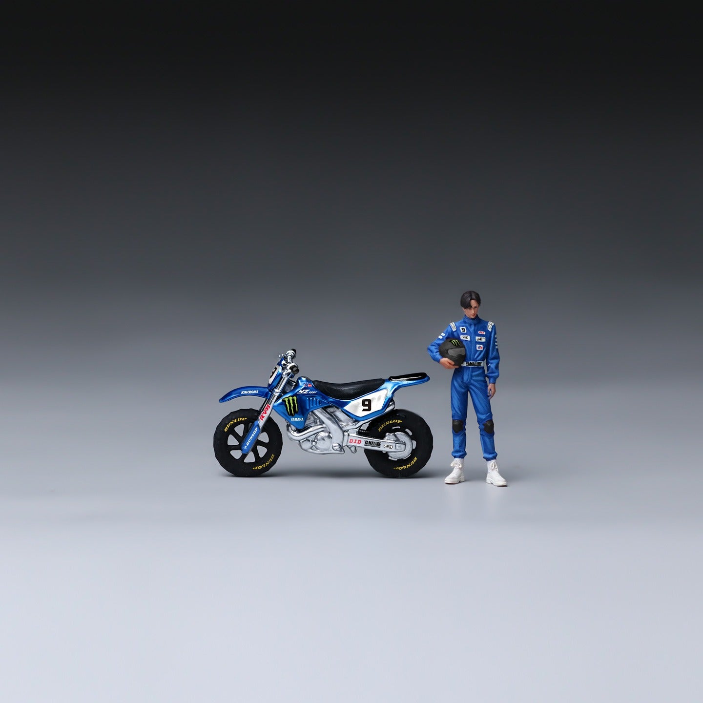 A miniature action figure of a race car driver in a blue racing suit standing next to a detailed blue and white dirt bike model, set against a dark-to-light gray studio gradient background.