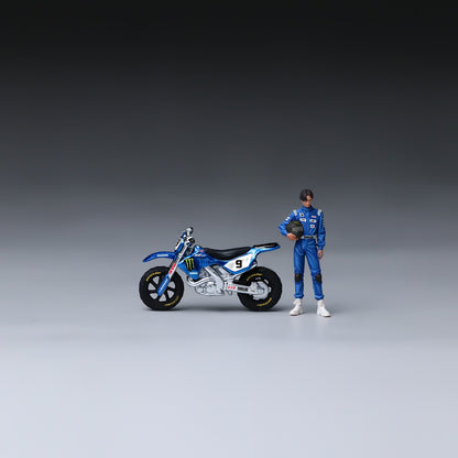 A miniature action figure of a race car driver in a blue racing suit standing next to a detailed blue and white dirt bike model, set against a dark-to-light gray studio gradient background.