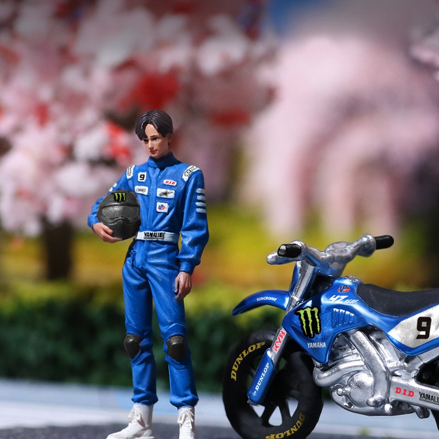 A detailed action figure of a male racer in a blue racing suit holding a helmet, standing next to a blue and white Yamaha dirt bike with Monster Energy and Dunlop logos, set against a blurred background of pink cherry blossoms.